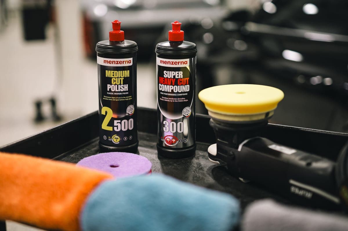 A bottle of compound and a cutting pad on a bright bench beside a polisher