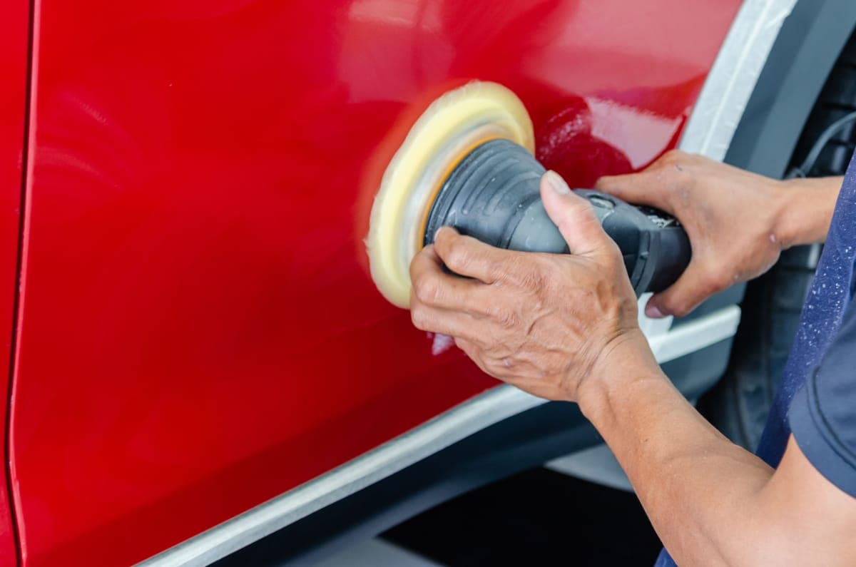 Two hands holding a polisher with a foam pad against a red car door