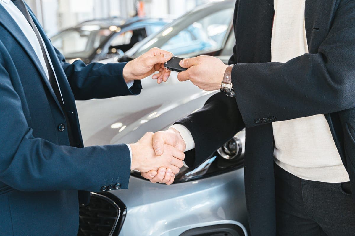 A car key changing hands in front of a car at a dealership