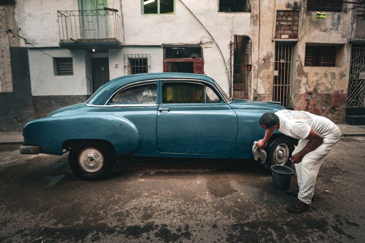 A man washing a classic car with a bucket and cloth on a street